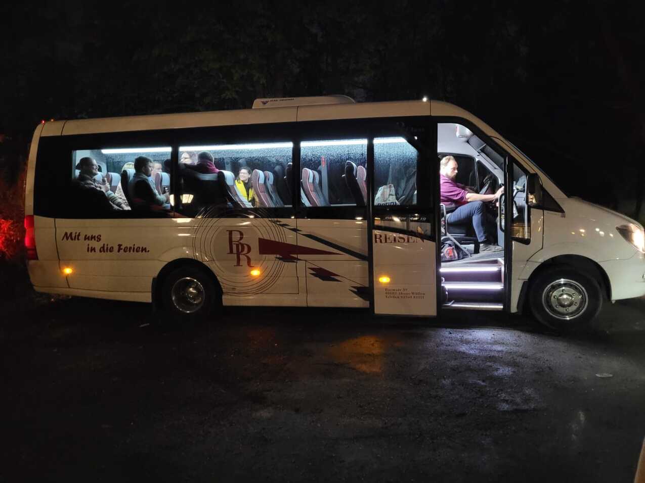 Nachtaufnahme eines Minibusses mit Passagieren, der an einer Haltestelle steht und von innen beleuchtet ist.