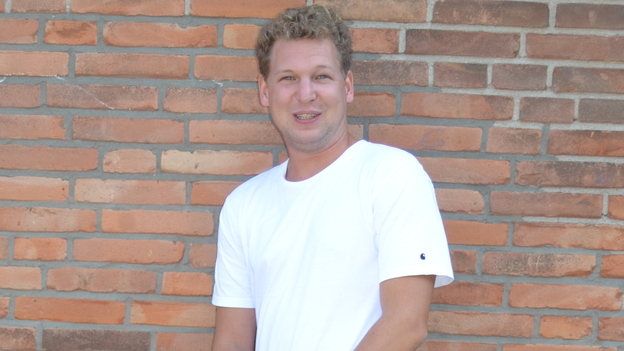 Junger Mann in wei&szlig;em T-Shirt und blauen Jeans steht vor einer ziegelsteinerne Wand.