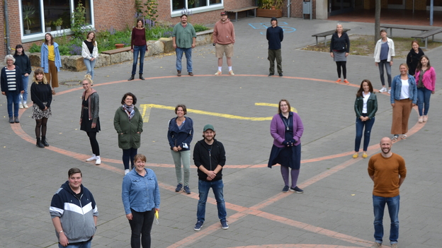 Gruppenfoto von Lehrkr&auml;ften im Schulhof, gleichm&auml;&szlig;ig verteilt in einem Kreis auf gepflastertem Boden.