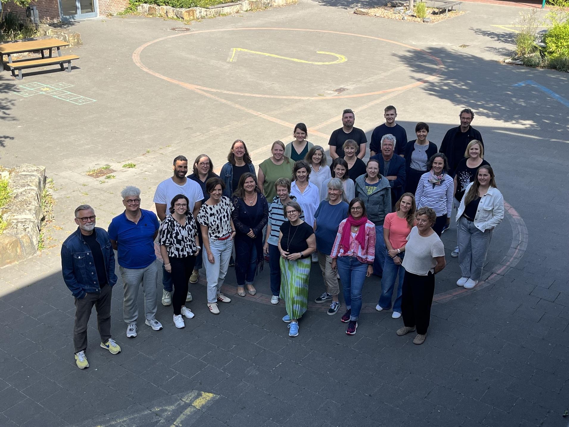 Gruppenfoto von 30 Personen auf einem Schulhof, umgeben von Bäumen, mit einer großen Zahl 2 im Hintergrund.