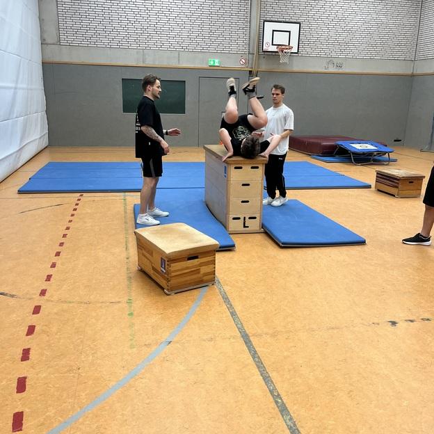 Drei Jugendliche trainieren in einer Turnhalle; einer springt &uuml;ber eine Box, w&auml;hrend die anderen unterst&uuml;tzen.