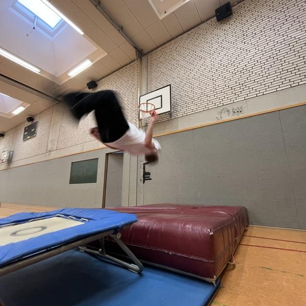 Ein Turner springt akrobatisch &uuml;ber ein Trampolin in einer Sporthalle mit Basketballkorb im Hintergrund.