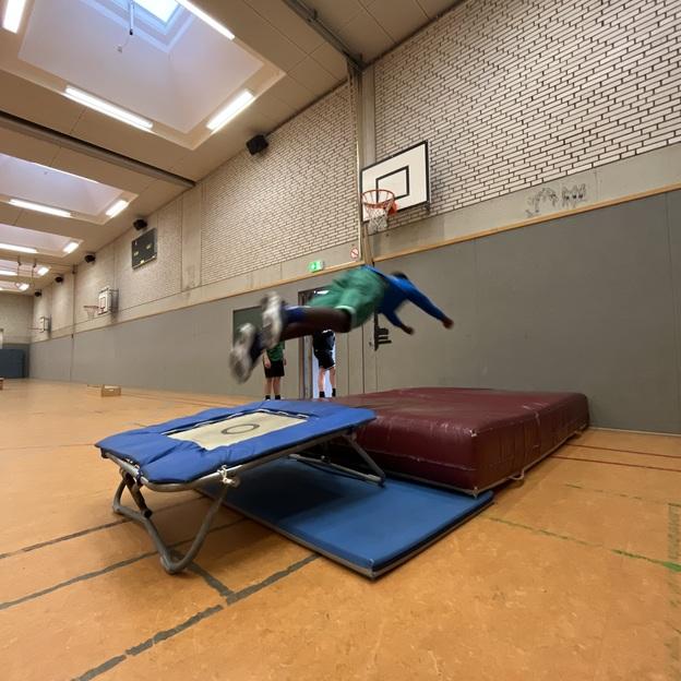 Junge springt von einem Trampolin auf eine Matte in einer Sporthalle. Basketballkorb im Hintergrund sichtbar.