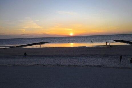 Sonnenuntergang am Strand mit Menschen, die sich am Wasser und auf dem Sand aufhalten, und einer ruhigen Meeresoberfläche.