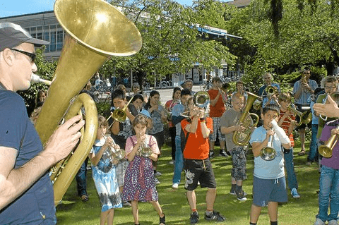 Ein Musiker mit Tuba leitet eine Gruppe von Kindern, die verschiedene Blechblasinstrumente im Freien spielen.