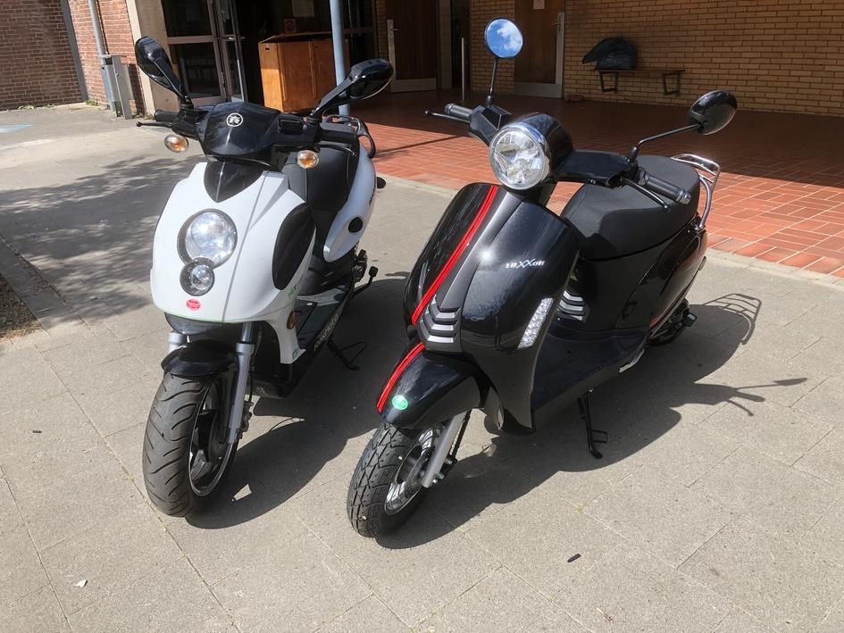 Zwei Motorroller, einer weiß und der andere schwarz, stehen auf einem grauen Pflaster vor einem Gebäude.