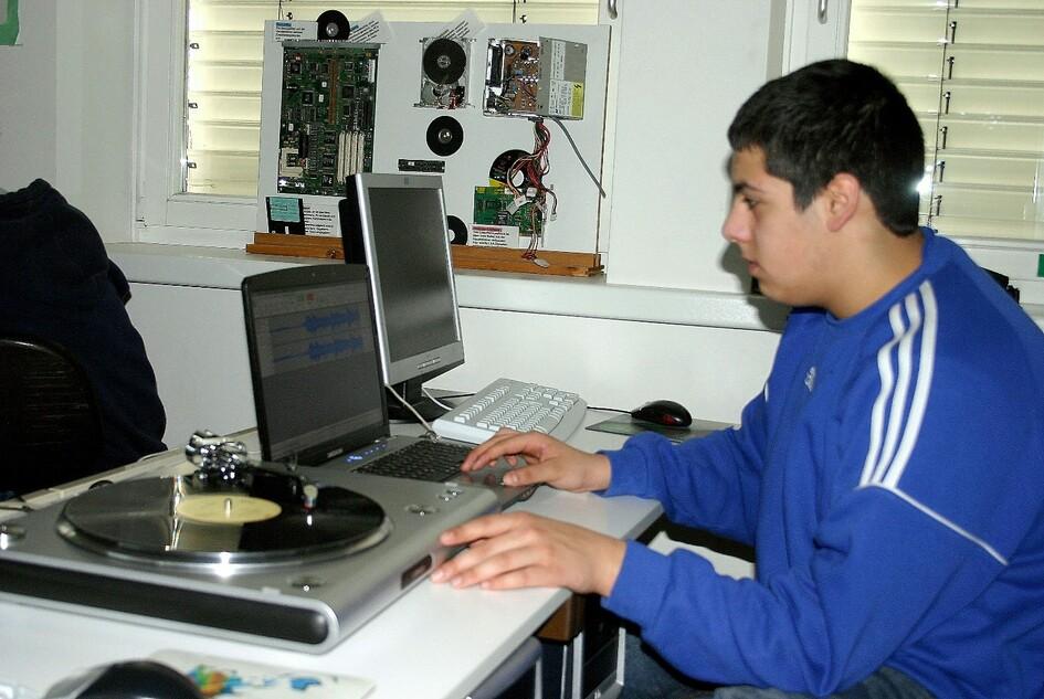 Junger Mann mit blauer Jacke arbeitet an einem Laptop neben einem Plattenspieler in einem Klassenraum.