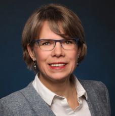 Porträt einer Frau mit kurzen Haaren, Brille und silbernen Ohrringen, lächelnd vor einem dunklen Hintergrund.