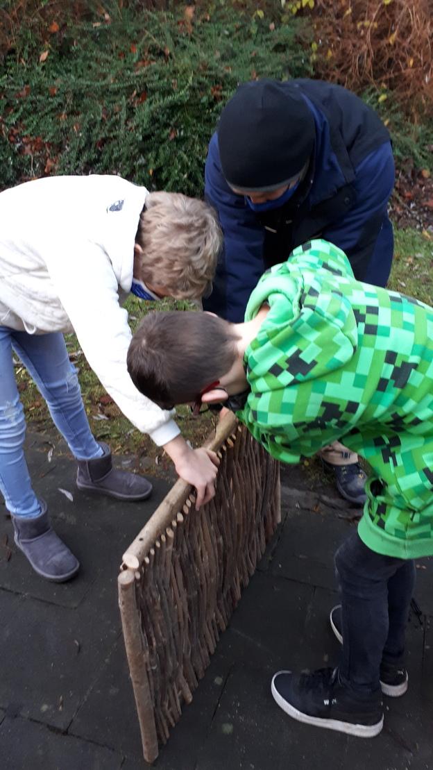 Drei Kinder arbeiten gemeinsam an einem Gestell aus Ästen auf dem Boden, umgeben von grünen Pflanzen.