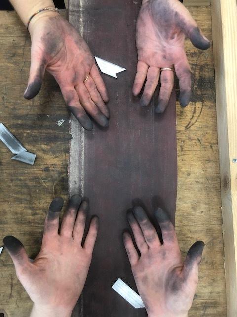 Vier H&auml;nde mit schwarzem Schmutz auf einer langen Holzlatte, umgeben von Werkzeugst&uuml;cken auf einem Holztisch.