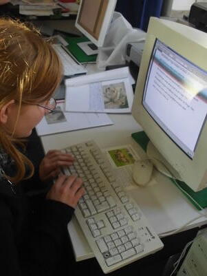 Sch&uuml;lerin tippt konzentriert auf einer Computertastatur vor einem Monitor, umgeben von Unterlagen.