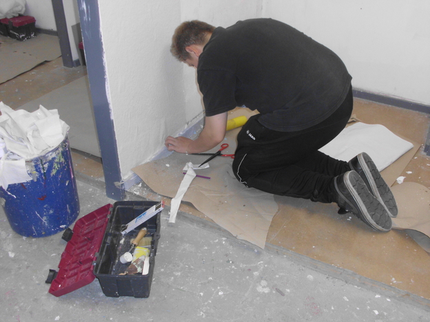 Junger Mann kniet, schneidet Material und arbeitet an einer Wand mit Werkzeugkiste und Farbtuben auf dem Boden.