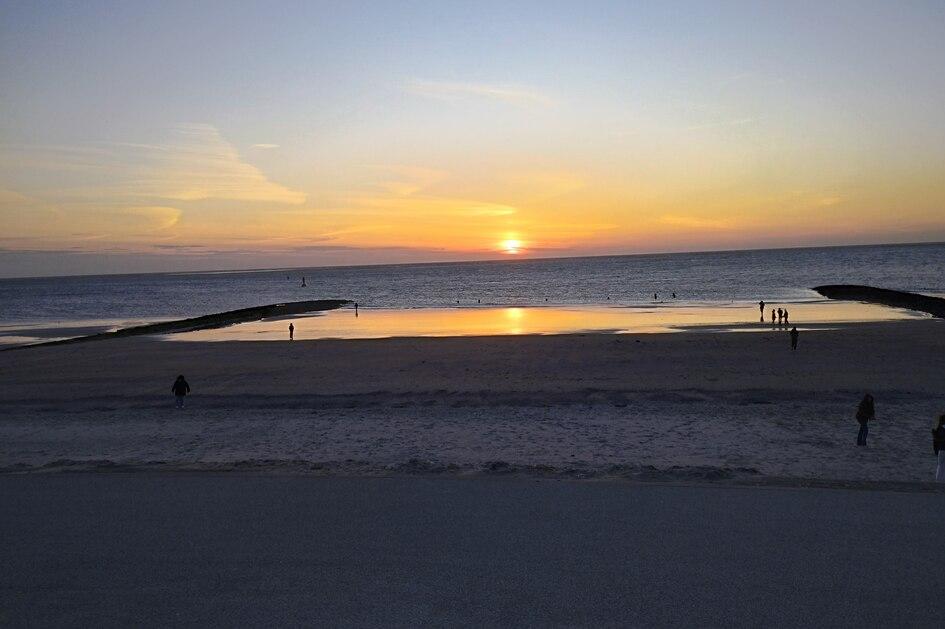 Sonnenuntergang am Strand mit Menschen, die sich am Wasser und auf dem Sand aufhalten, und einer ruhigen Meeresoberfläche.