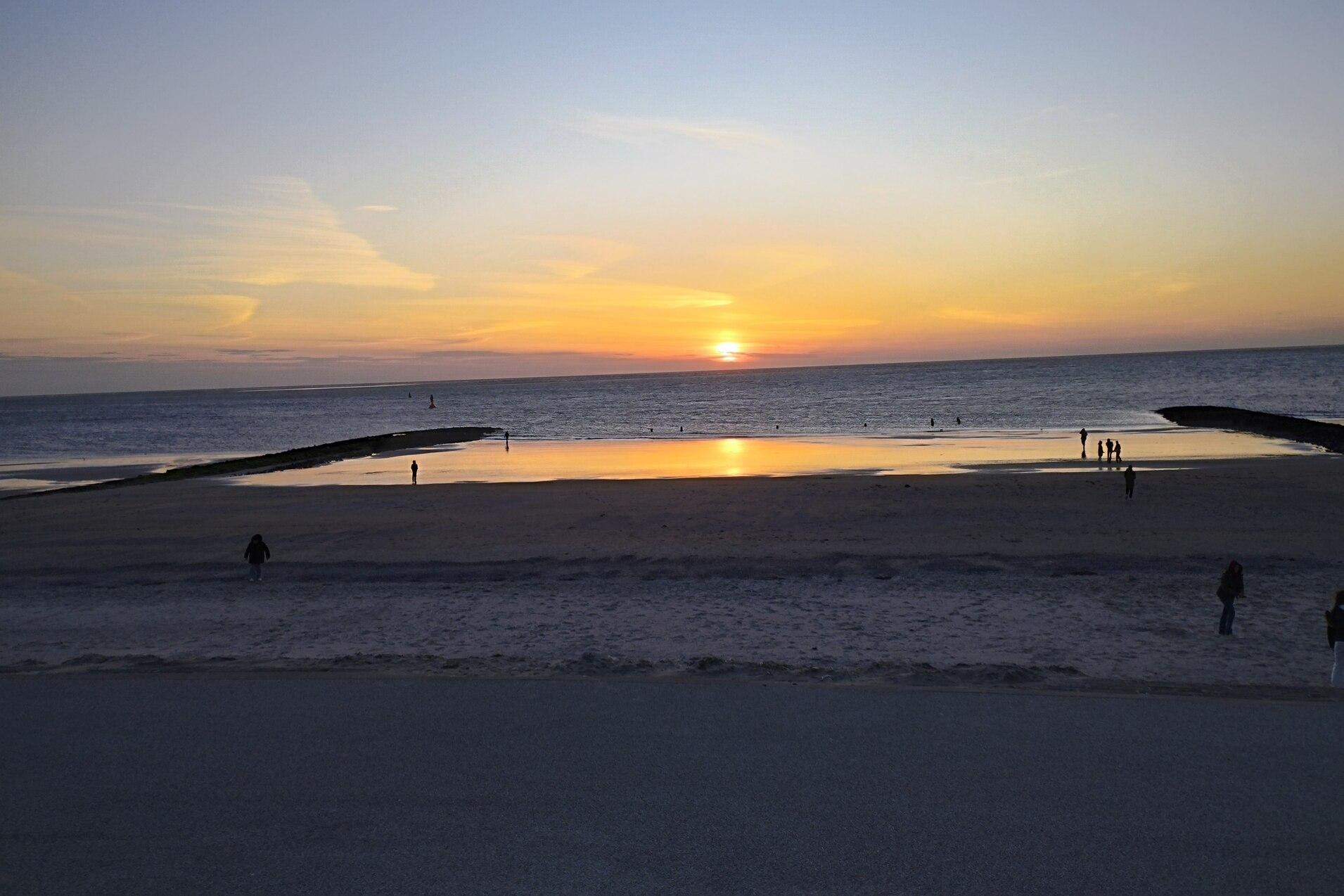 Sonnenuntergang am Strand mit Menschen, die sich am Wasser und auf dem Sand aufhalten, und einer ruhigen Meeresoberfl&auml;che.