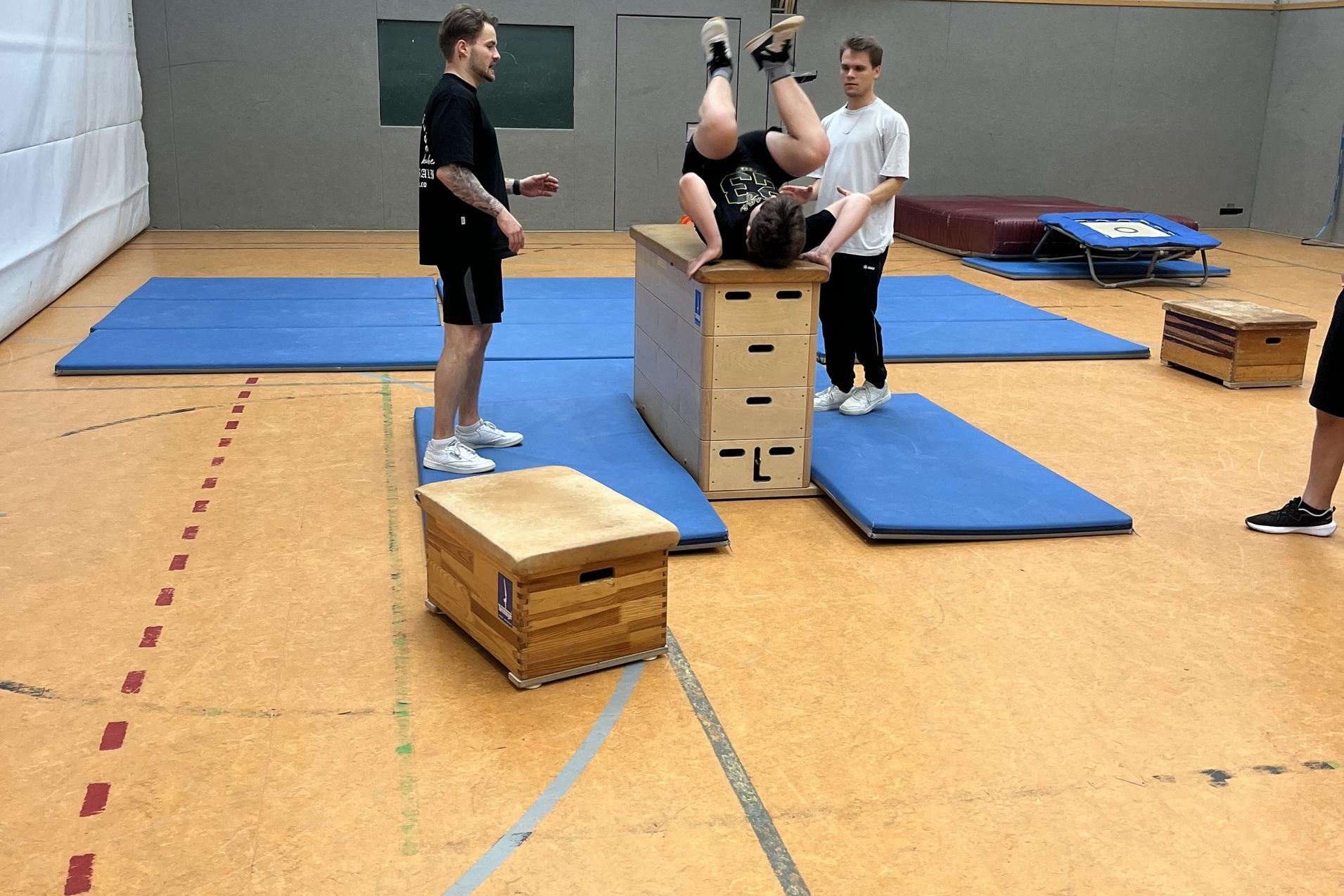 Drei Jugendliche trainieren in einer Turnhalle; einer springt &uuml;ber eine Box, w&auml;hrend die anderen unterst&uuml;tzen.