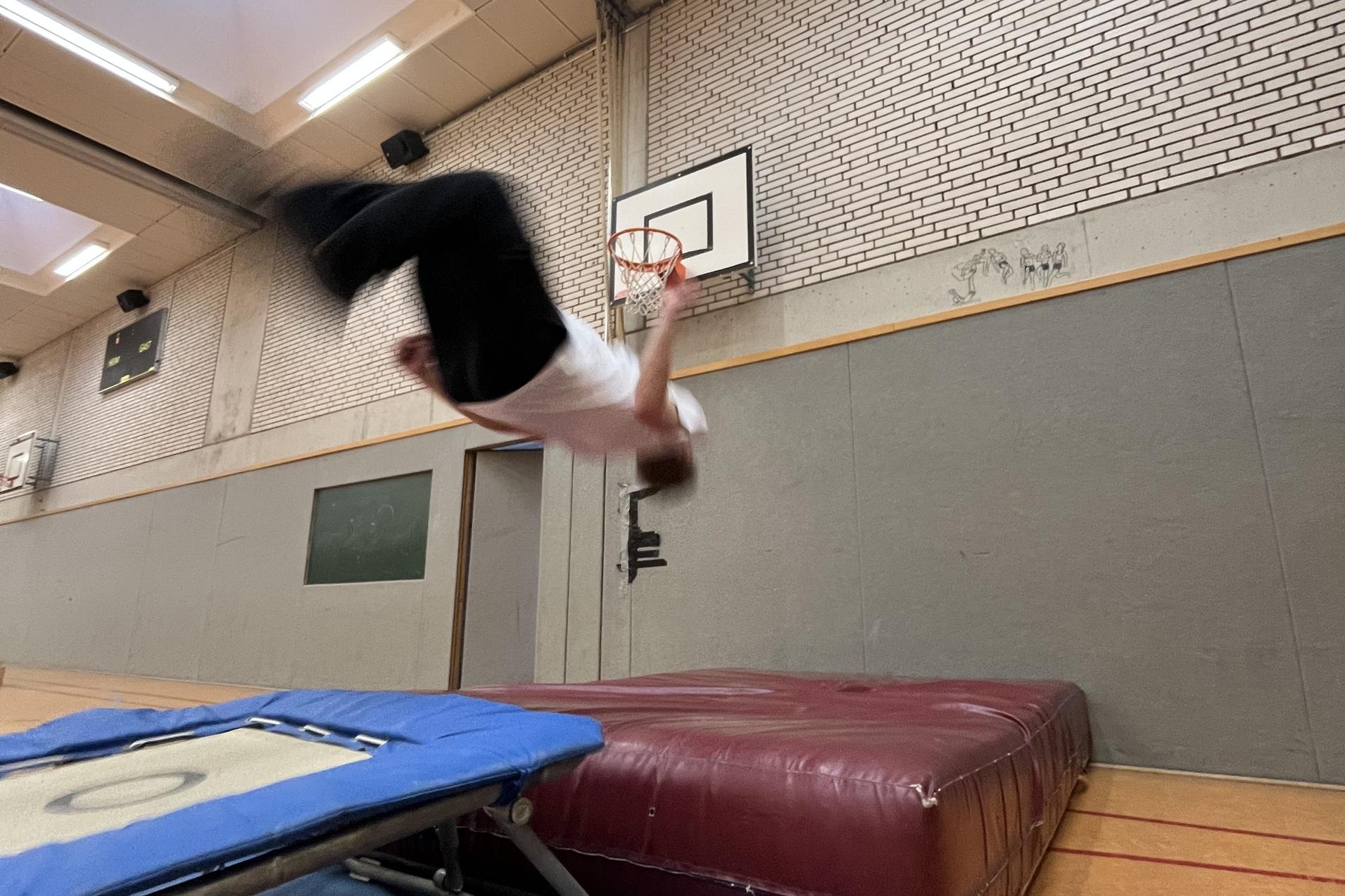 Ein Turner springt akrobatisch &uuml;ber ein Trampolin in einer Sporthalle mit Basketballkorb im Hintergrund.