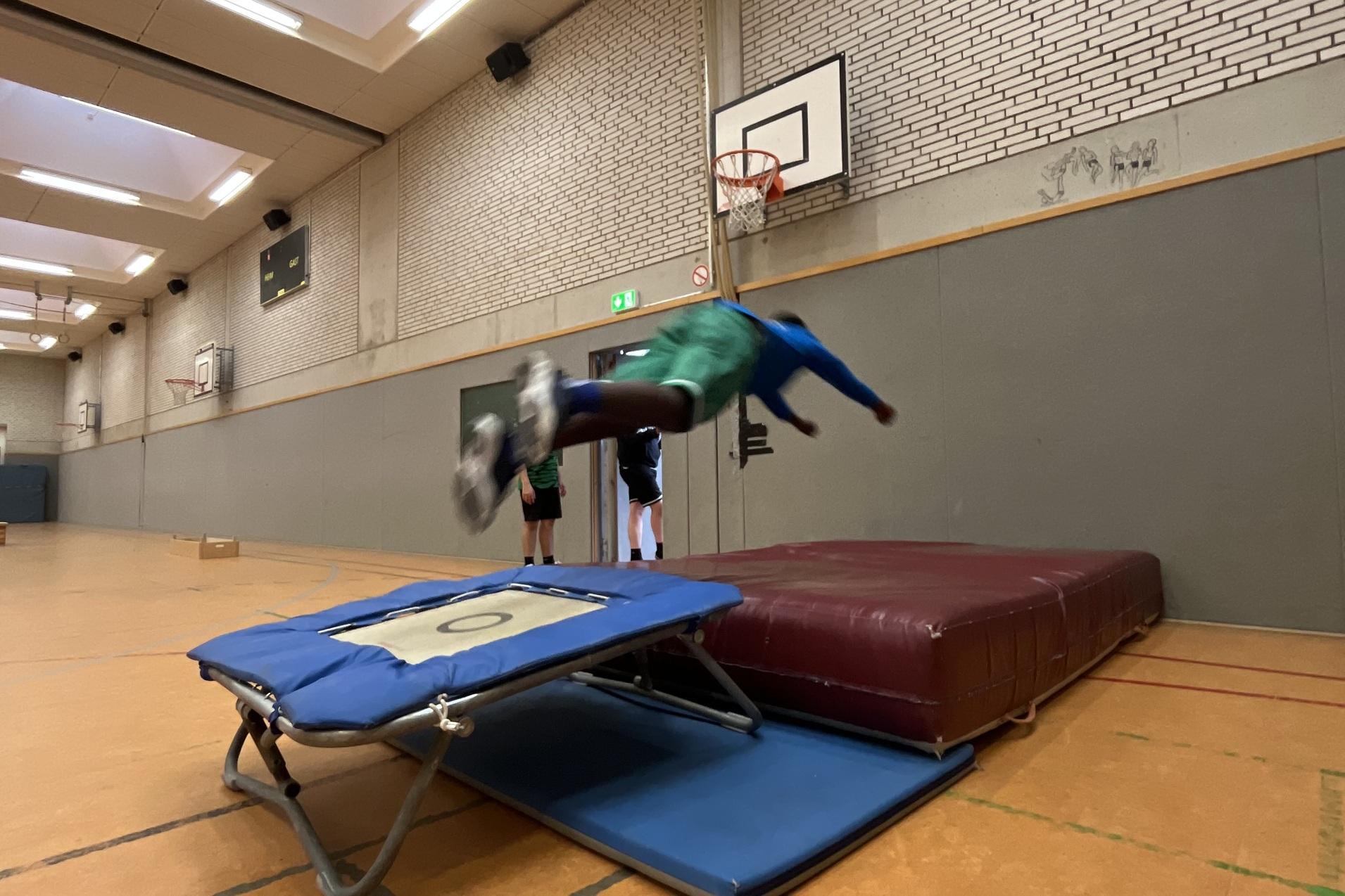 Junge springt von einem Trampolin auf eine Matte in einer Sporthalle. Basketballkorb im Hintergrund sichtbar.