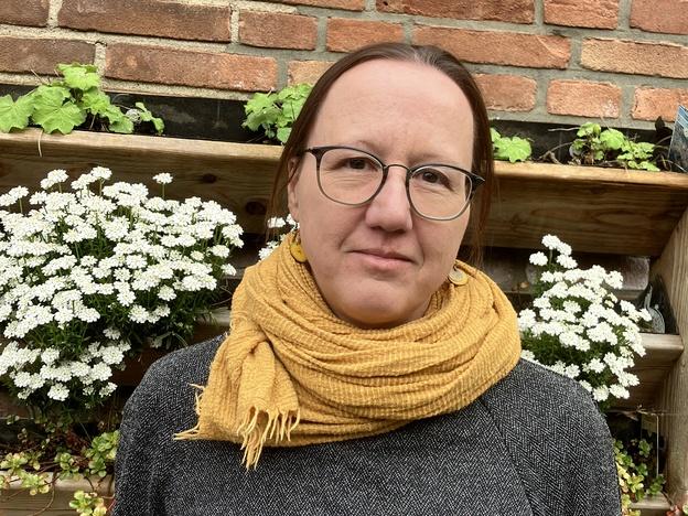 Frau mit Brille und gelbem Schal vor einer Holzstruktur mit blühenden Pflanzen und einer Ziegelfassade im Hintergrund.