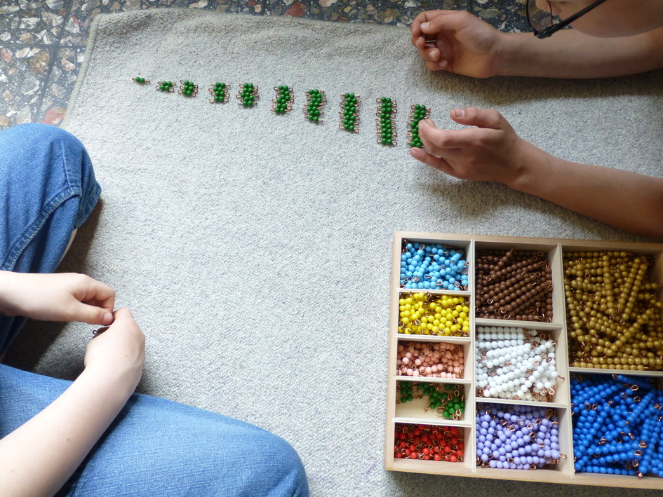 Zwei H&auml;nde sortieren bunte Perlen auf einem Teppich, w&auml;hrend eine Schachtel mit weiteren Perlen daneben steht.
